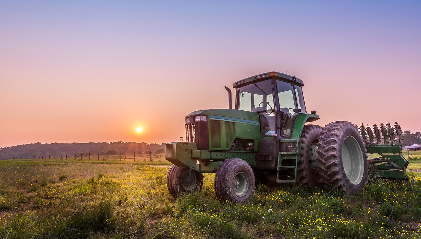 Tractor verde con implemento agrícola estacionado en campo de pasto al atardecer, con cielo en tonos cálidos naranjas y púrpuras