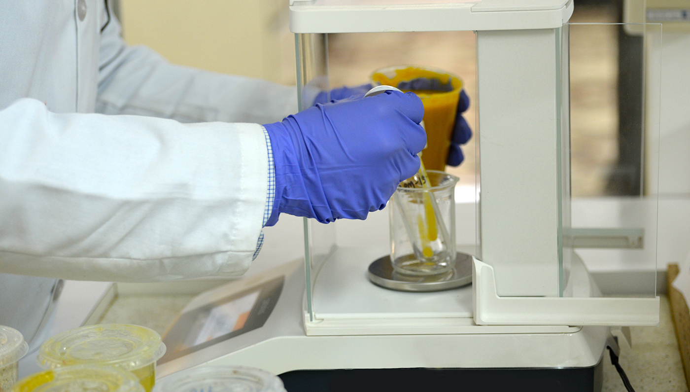 Primer plano de una mano con guante colocando un recipiente con material amarillo sobre una báscula de laboratorio, rodeada de matraces y equipo, con tonos claros y ambiente científico.