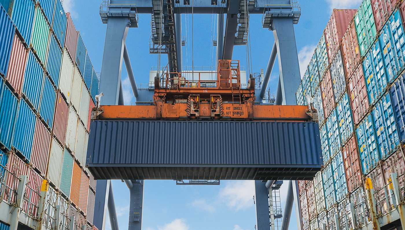 Vista amplia de una grúa pórtico naranja levantando contenedores de carga entre torres multicolores bajo un cielo azul, con tonos cálidos y ambiente logístico industrial.